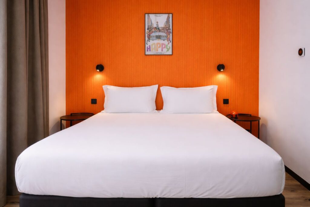 Grand lit blanc avec tête de lit orange vif et affiche "Happy", chambre double du ClapClap Hotel Strasbourg