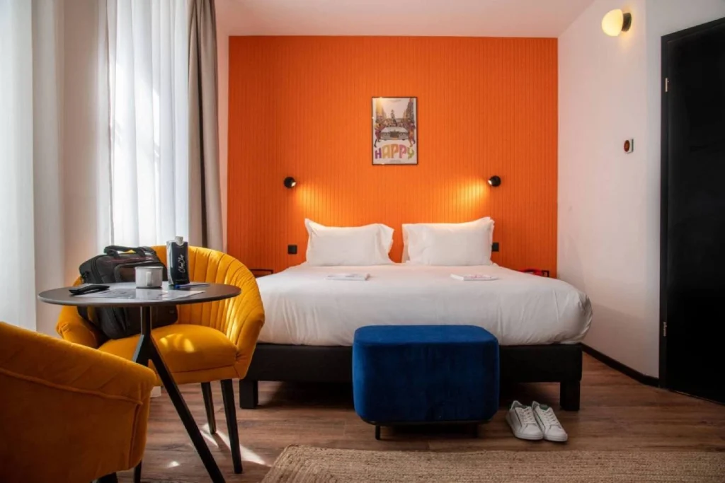Chambre Double Supérieure du ClapClap Hôtel Strasbourg avec lit 180x200, mur orange, fauteuils jaunes, pouf bleu et affiche "Happy"