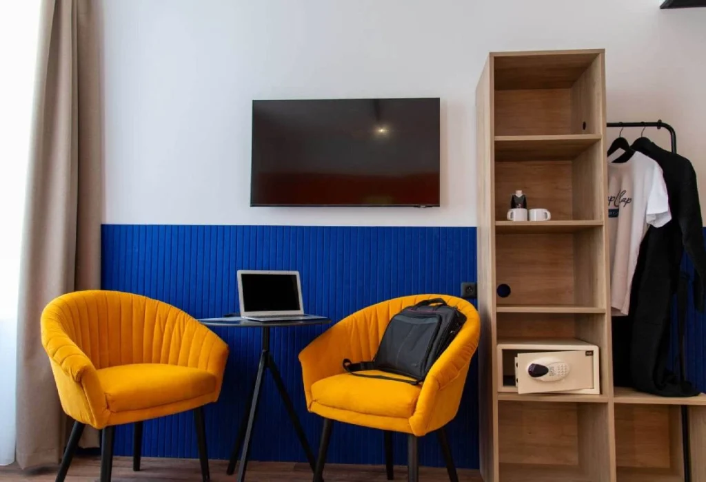 Chambre Double Supérieure du ClapClap Hôtel Strasbourg avec deux fauteuils velours jaune, TV murale, mur bleu, étagère bois, coffre-fort et penderie