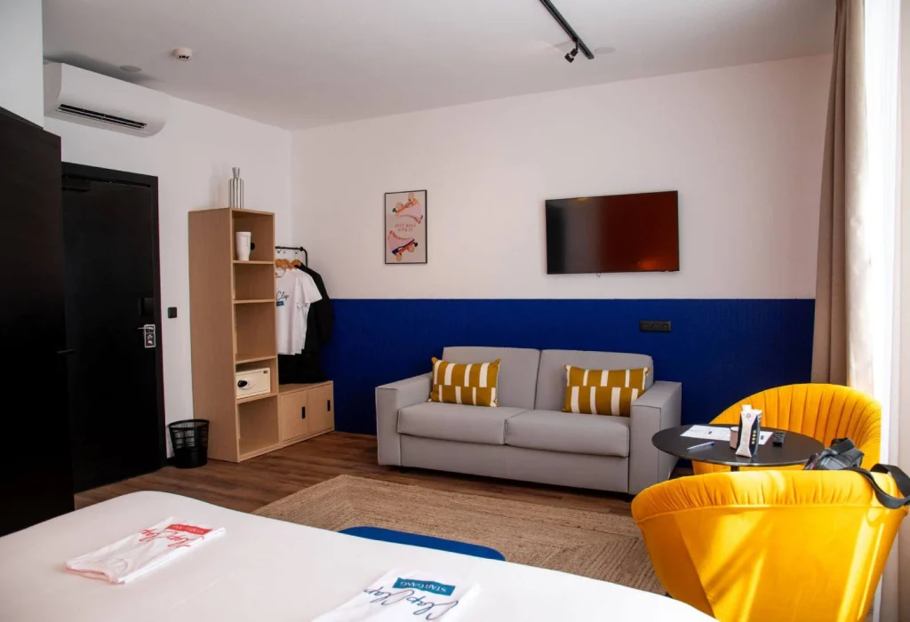 Chambre Triple du ClapClap Hôtel Strasbourg avec lit, canapé gris, fauteuil jaune, mur bleu, TV murale et étagère bois