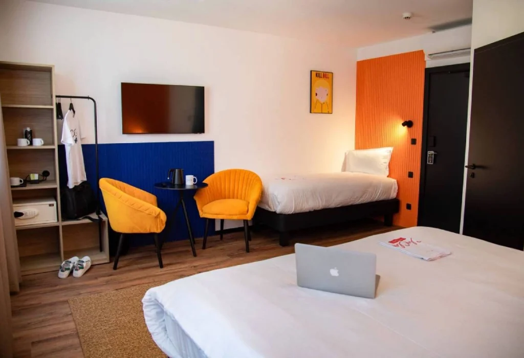 Chambre Triple du ClapClap Hôtel Strasbourg avec deux lits, fauteuils jaunes, murs bleu et orange, TV murale et étagère bois