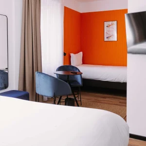 Chambre Triple du ClapClap Hôtel Strasbourg avec deux lits, mur orange, chaises bleues et table ronde
