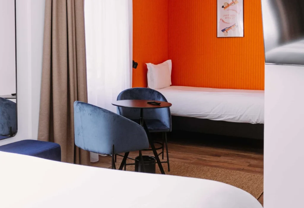 Chambre Triple du ClapClap Hôtel Strasbourg avec deux lits, mur orange, chaises bleues et table ronde