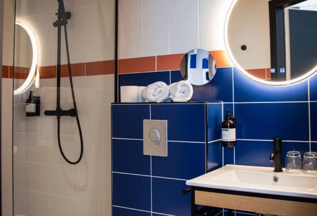 Salle de bain de la Chambre Double Supérieure du ClapClap Hôtel Strasbourg avec douche vitrée, carrelage bleu, deux miroirs ronds rétroéclairés et vasque
