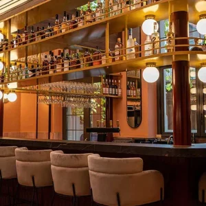 Comptoir du bar Clap Show Strasbourg avec tabourets beige, étagères de spiritueux et éclairage globe doré