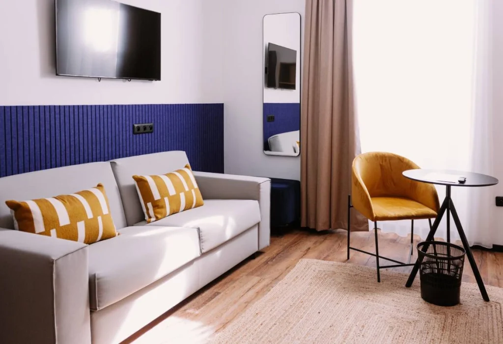 Chambre Quadruple du ClapClap Hôtel Strasbourg avec canapé gris, coussins ocre, mur bleu, TV murale et fauteuil ocre