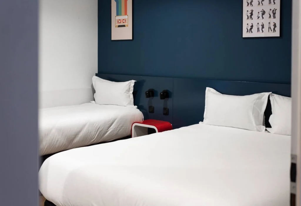 Chambre Tribu du ClapClap Hôtel Strasbourg avec deux lits, mur bleu pétrole, tabouret rouge et affiches murales